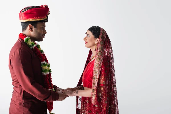 Vista lateral de la feliz pareja india de marido y mujer en ropa tradicional cogidos de la mano aislados en gris - foto de stock