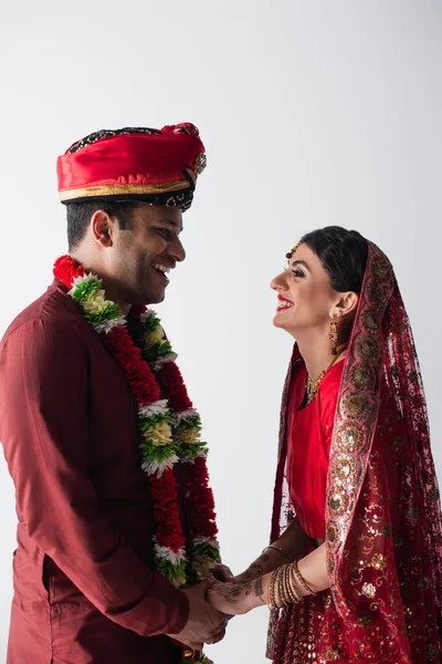 Vista lateral de alegre marido indio y esposa en ropa tradicional tomados de las manos aislados en blanco - foto de stock
