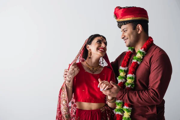 Веселый индийский муж и жена в традиционной одежде держась за руки изолированные на сером — стоковое фото