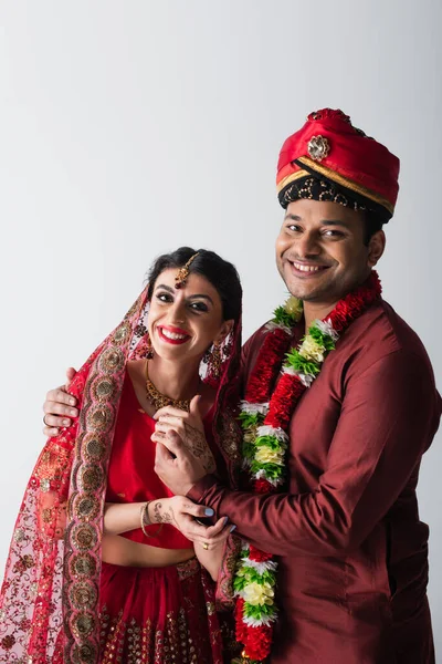 Marido y mujer indios positivos en ropa tradicional tomados de las manos aislados en gris - foto de stock