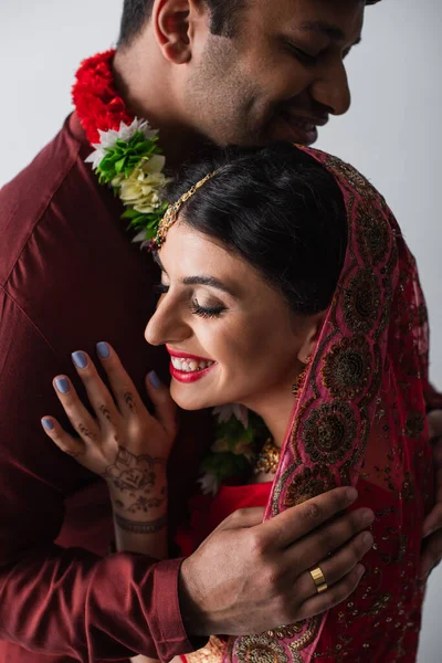 Bridegroom hugging pleased indian bride in headscarf isolated on grey — Stock Photo