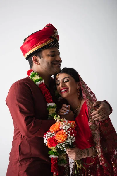 Счастливый индиец в тюрбане обнимает веселую невесту в сари с цветами, изолированными на сером — стоковое фото