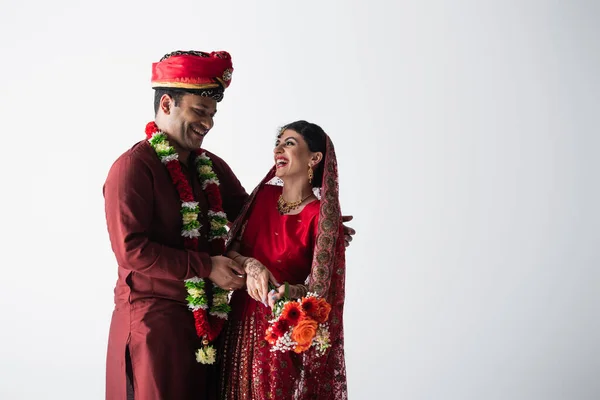Счастливый индиец в тюрбане смотрит на веселую невесту в сари с букетом цветов, изолированных на сером — стоковое фото