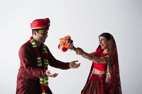 Homme indien en turban attrapant bouquet de fleurs près de la mariée gaie en sari isolé sur gris — Photo de stock