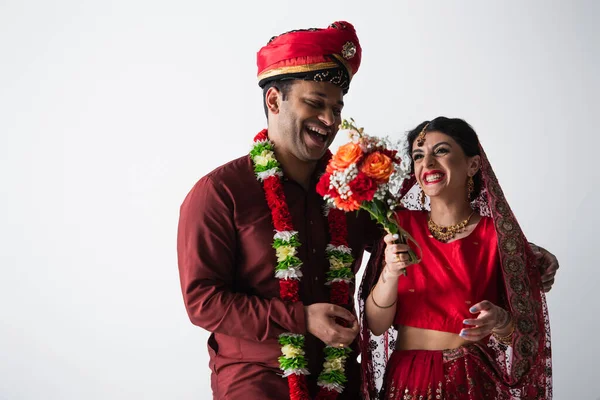 Веселый индийский мужчина в тюрбане смотрит на невесту в сари с букетом цветов, изолированных на сером — стоковое фото