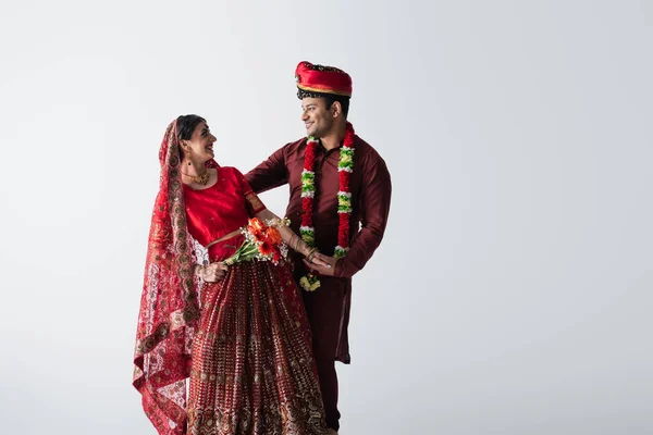 Улыбающийся индиец в тюрбане смотрит на красивую невесту в сари с букетом цветов, изолированных на белом — стоковое фото