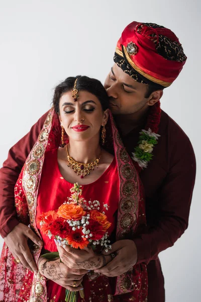 Hombre indio en turbante oliendo pelo de novia bonita en sari con ramo de flores aisladas en gris - foto de stock