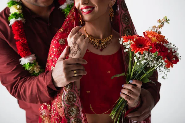 Vue recadrée de l'homme indien avec anneau sur le doigt tenant la main de mariée heureuse avec bouquet de fleurs isolées sur blanc — Photo de stock