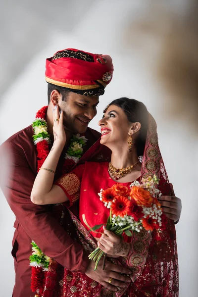 Homem indiano feliz abraçando noiva alegre em lenço de cabeça e sari com buquê de flores em branco — Fotografia de Stock