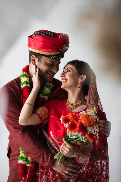 Heureux homme indien étreignant mariée gaie en foulard et sari avec bouquet de fleurs sur blanc — Photo de stock