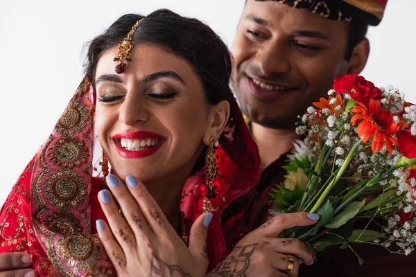 Hombre indio borroso en turbante mirando a la novia sonriente con mehndi sosteniendo flores en blanco - foto de stock