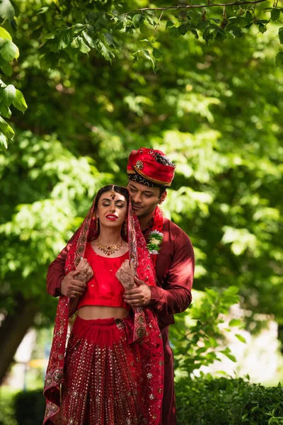 Hombre indio feliz en turbante abrazando novia con mehndi en sari y pañuelo en la cabeza - foto de stock