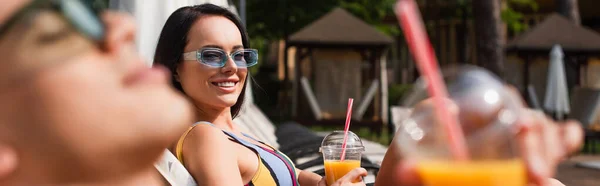 Mujer sonriente en gafas de sol mirando borrosa novio con jugo de naranja al aire libre, pancarta - foto de stock