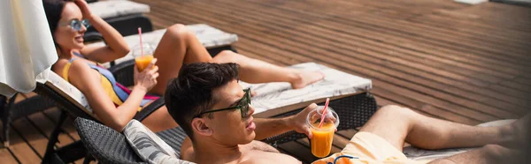 Человек в солнцезащитных очках держит апельсиновый сок рядом с размытой девушкой на шезлонге, баннер — стоковое фото