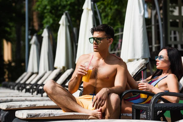 Pareja joven en traje de baño sosteniendo jugo de naranja en la silla de cubierta - foto de stock