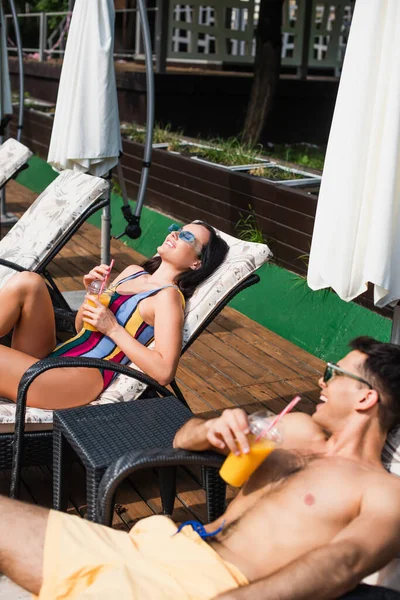 Vista de alto ângulo da mulher alegre em roupa de banho deitada com os olhos fechados na cadeira de praia perto do namorado com suco de laranja — Fotografia de Stock