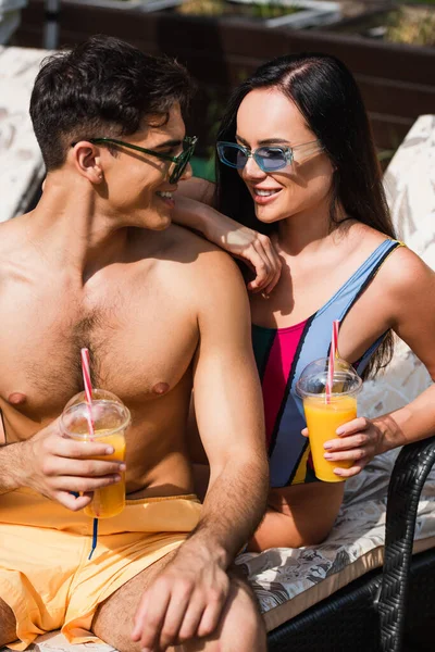 Un homme souriant dans des maillots de bain regardant sa petite amie avec du jus d'orange sur une chaise longue — Photo de stock