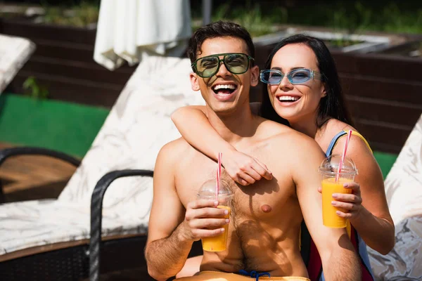 Mujer feliz con jugo de naranja abrazando novio en gafas de sol en la silla de cubierta - foto de stock