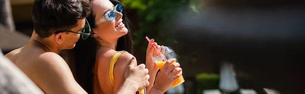 Hombre sin camisa abrazando novia sonriente con jugo de naranja al aire libre, pancarta - foto de stock