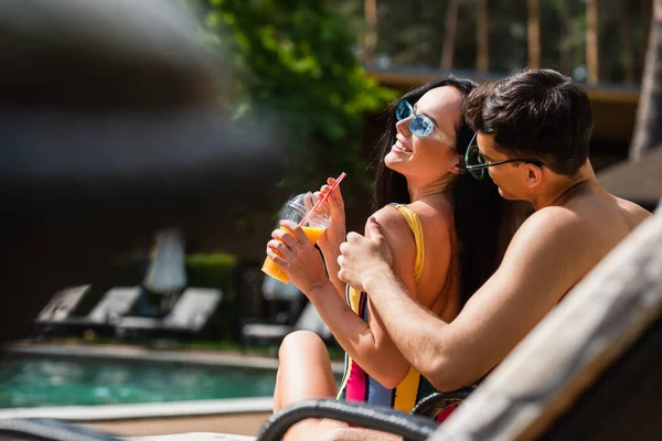 Junger Mann umarmt fröhliche Freundin im Resort mit Orangensaft — Stockfoto