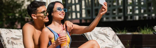 Happy couple with orange juice taking selfie on deck chair, banner — Stock Photo