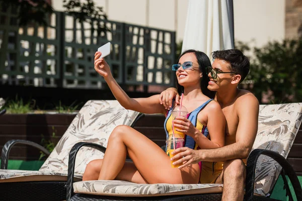 Young shirtless man hugging girlfriend with orange juice taking selfie on deck chair — Stock Photo