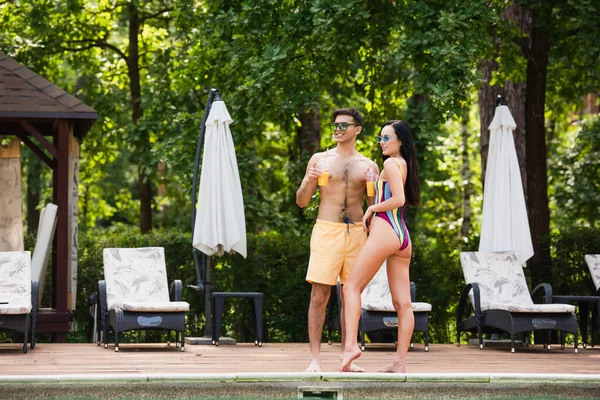 Mujer en traje de baño sosteniendo jugo de naranja cerca de novio y piscina - foto de stock