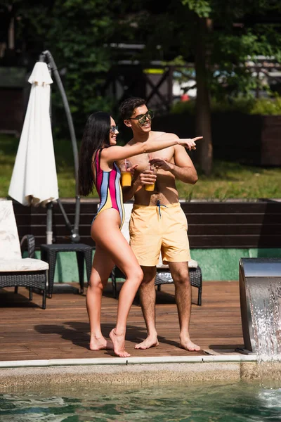 Mulher alegre em maiô apontando com o dedo perto do namorado e piscina — Fotografia de Stock