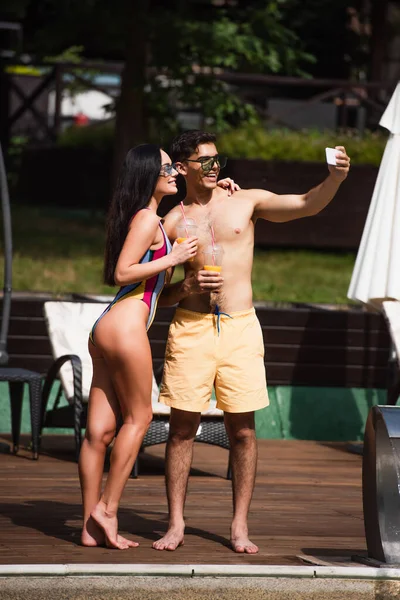 Smiling man with orange juice taking selfie on smartphone near girlfriend on resort — Stock Photo