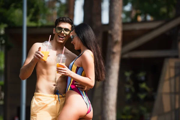 Shirtless man with orange juice hugging girlfriend and pointing with finger on resort — Stock Photo