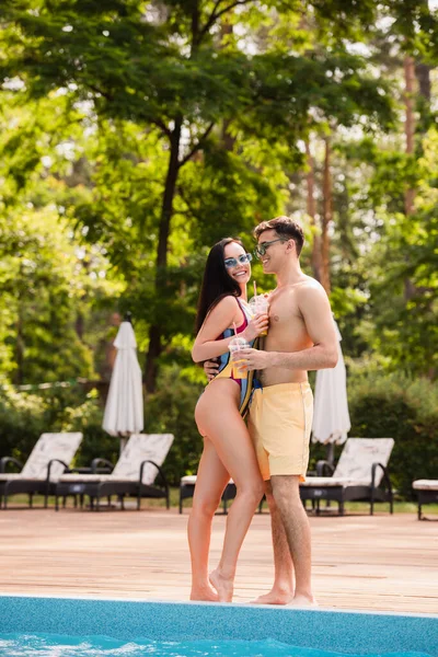 Hombre sin camisa abrazando novia sonriente con jugo de naranja cerca de la piscina - foto de stock