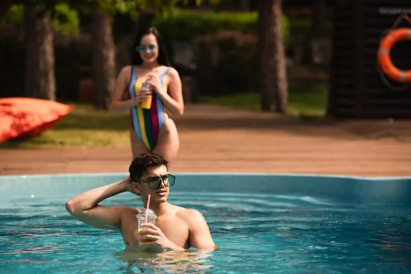 Jovem de óculos de sol segurando suco de laranja perto da namorada no fundo borrado na piscina — Fotografia de Stock