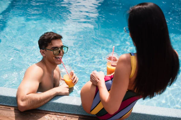 Улыбающийся мужчина пьет апельсиновый сок возле размытой женщины в бассейне — стоковое фото