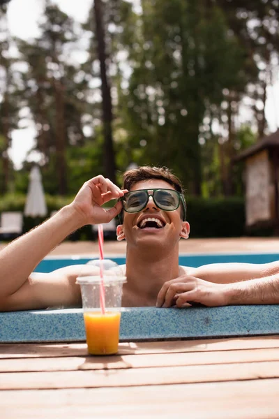 Uomo positivo che tiene gli occhiali da sole vicino al succo d'arancia sfocato a bordo piscina — Foto stock