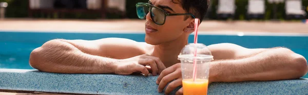 Homem de óculos de sol relaxante na piscina perto de suco de laranja borrado, banner — Fotografia de Stock