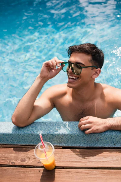 Vista de ángulo alto del hombre en gafas de sol sonriendo mientras se relaja en la piscina cerca del jugo de naranja - foto de stock