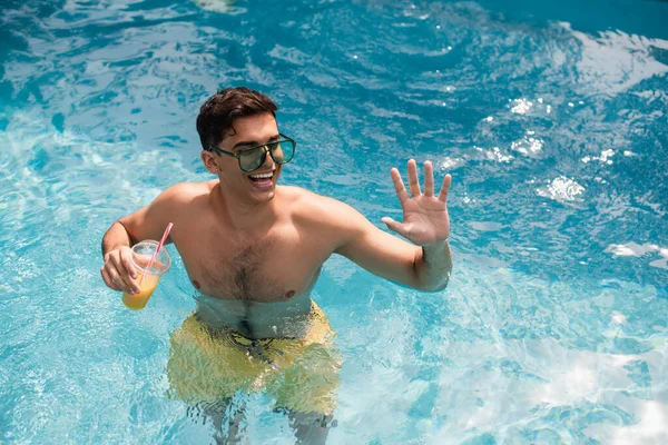 High angle view of smiling man with orange juice waving hand in swimming pool — Stock Photo
