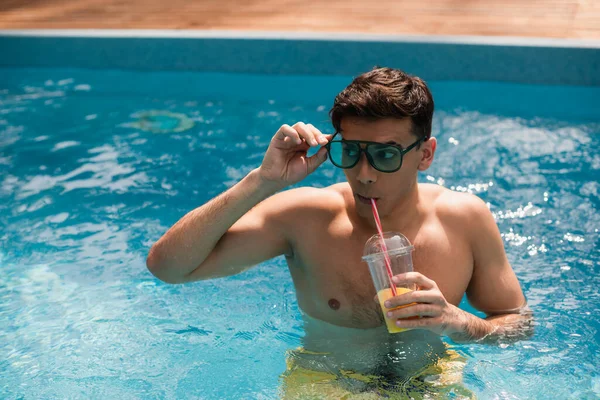 Uomo che regola gli occhiali da sole mentre beve succo d'arancia in piscina — Foto stock