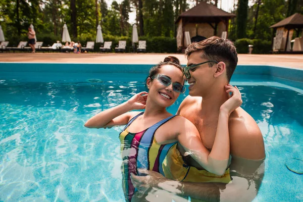 Hombre sonriente en gafas de sol abrazando novia con los ojos cerrados en la piscina - foto de stock