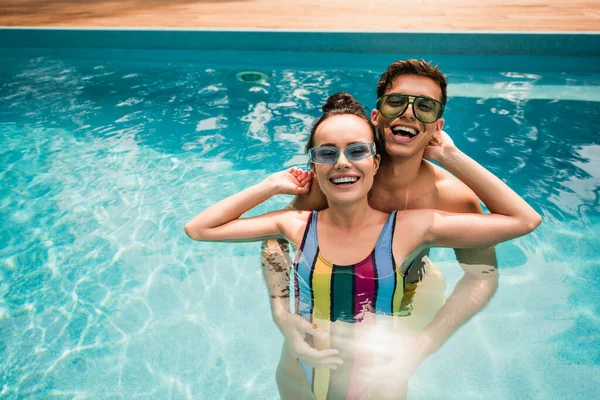 Coppia felice in occhiali da sole guardando la fotocamera mentre in piedi in piscina — Foto stock
