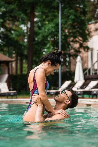 Seitenansicht positiver Mann steigt gut gelaunte Freundin im Urlaub im Schwimmbad auf — Stockfoto