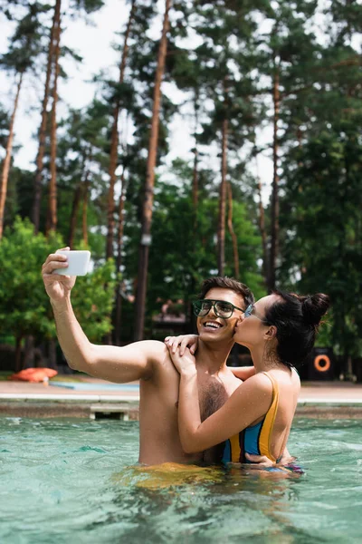 Young woman kissing boyfriend taking selfie on mobile phone in swimming pool — Stock Photo