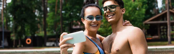 Smiling woman in swimsuit taking selfie on blurred smartphone and hugging boyfriend on resort, banner — Stock Photo