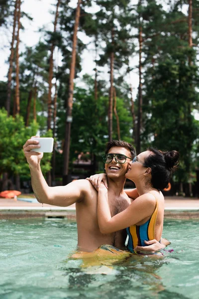 Pretty woman kissing and embracing boyfriend taking selfie in swimming pool — Stock Photo