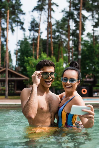 Pareja feliz en gafas de sol tomando selfie en smartphone en la piscina durante el fin de semana - foto de stock