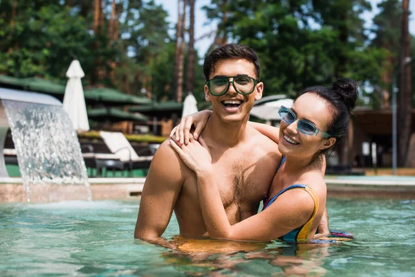 Casal alegre abraçando e olhando para a câmera na piscina — Fotografia de Stock