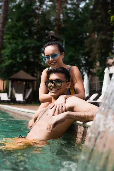 Femme souriante relaxant avec petit ami dans la piscine près fontaine floue — Photo de stock