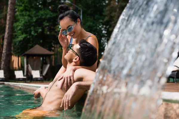 Donna che tiene gli occhiali da sole e abbraccia il fidanzato vicino alla fontana in piscina — Foto stock