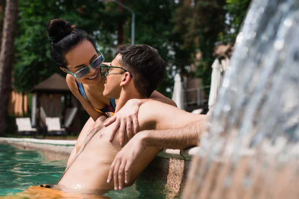 Jovem mulher sorrindo para namorado na piscina com fonte — Fotografia de Stock