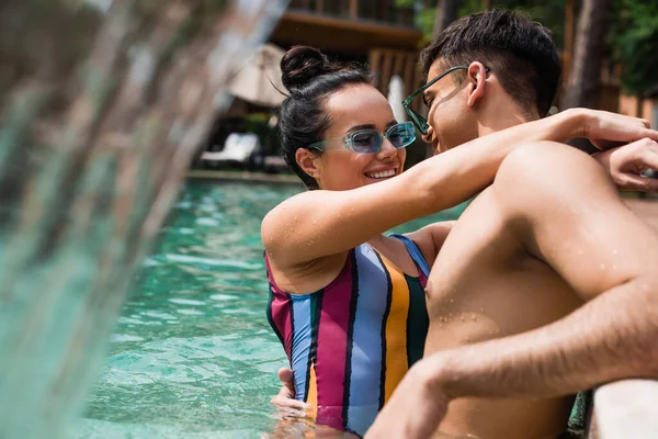 Jeune couple dans des lunettes de soleil étreignant près fontaine floue dans la piscine — Photo de stock